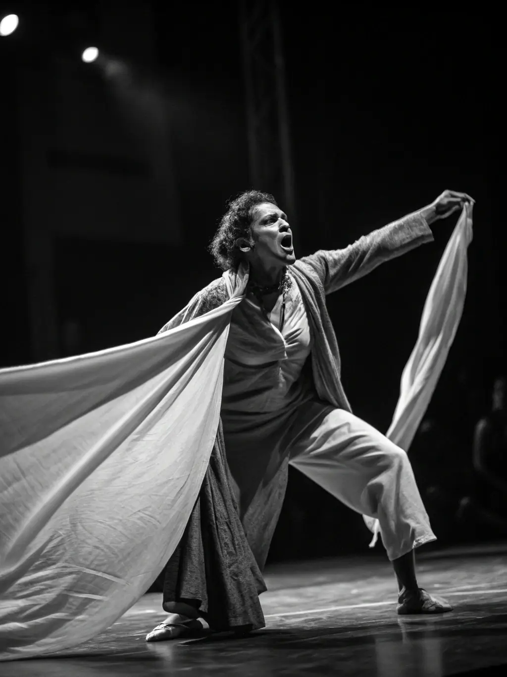 A black and white image showcasing a solo theatrical performance, with the artist in costume conveying a powerful emotion through their acting.